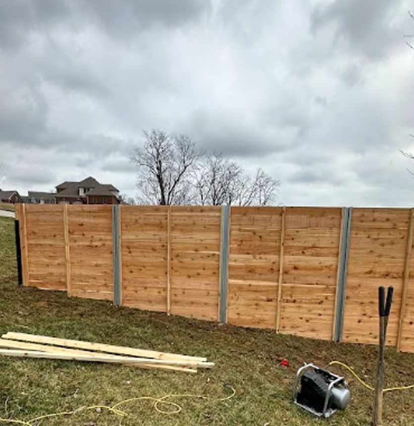 Wooden backyard fence panels with concrete posts under a cloudy sky, with lumber and tools in the grass.