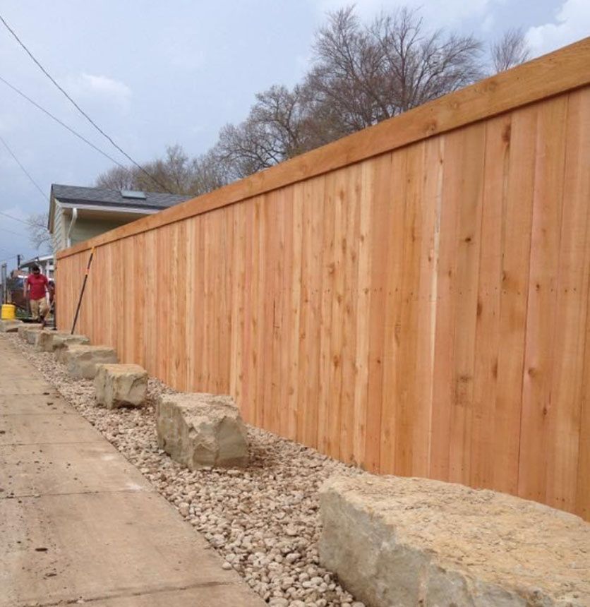Fresh wooden privacy fence along a sidewalk, with rocks and concrete blocks in front under cloudy sky