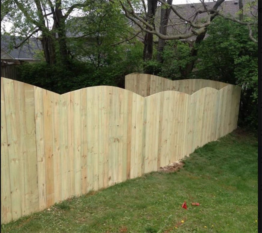New wooden fence with wavy top along a grassy yard, with trees and houses behind it