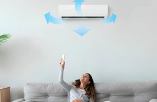 A woman is sitting on a couch and holding a remote control, pointing it at the air conditioner.