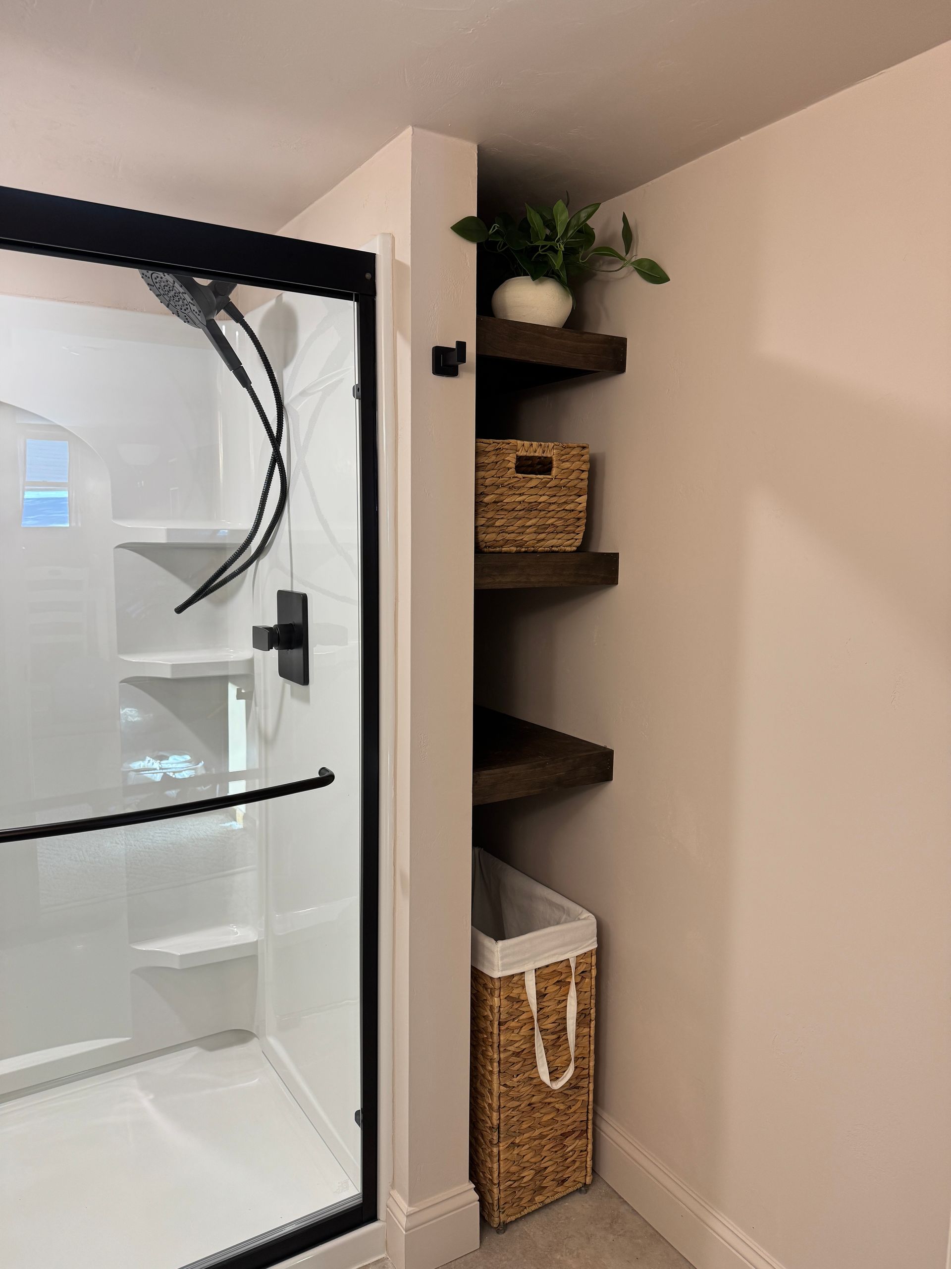 Bathroom corner with dark shelves, white shower, and woven basket. A plant sits on the top shelf.