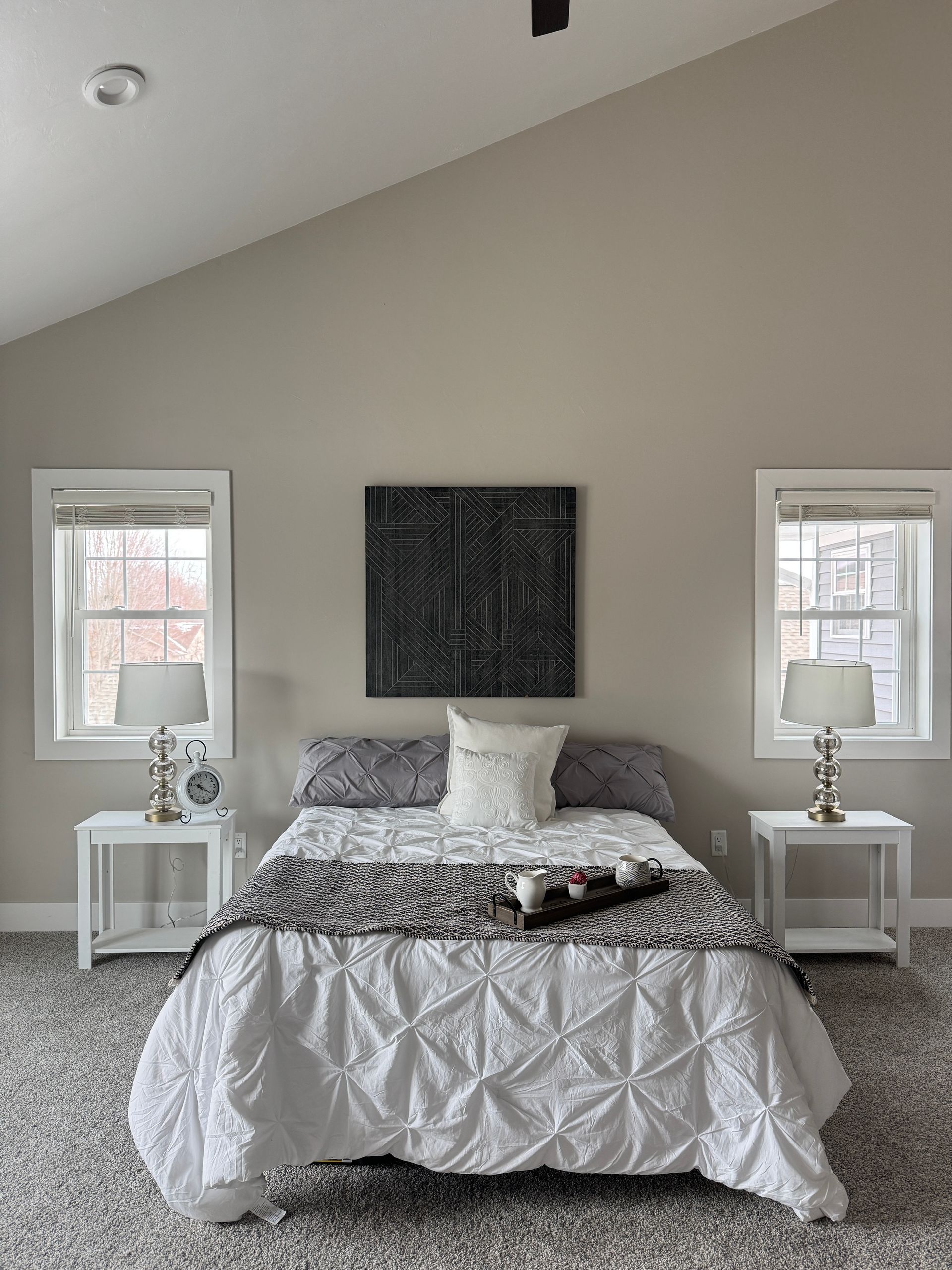 Bedroom with bed, nightstands, windows, and artwork on a neutral-colored wall.