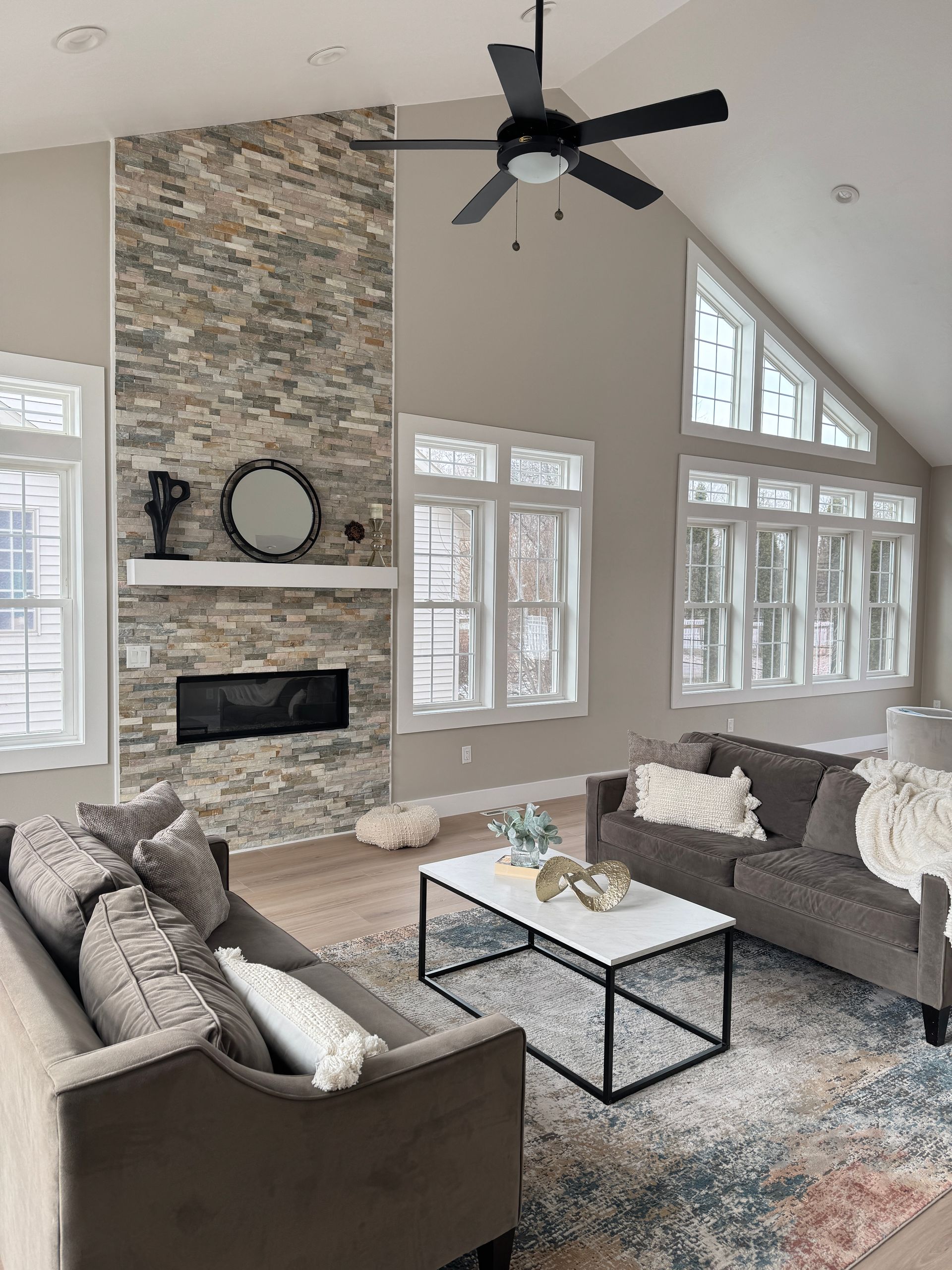 Living room with stone fireplace, gray couches, and a black ceiling fan.