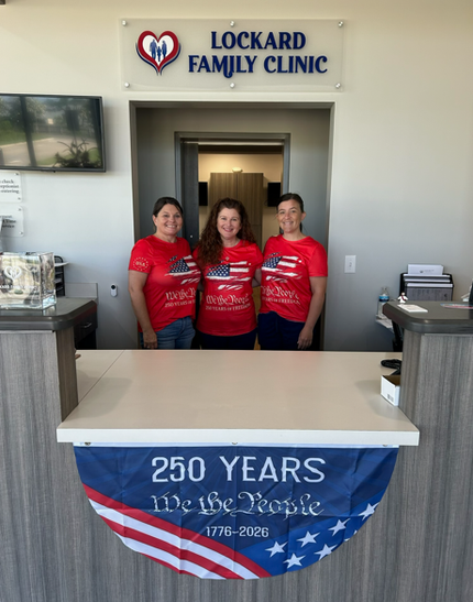 Three women in red shirts stand at a clinic desk with a “250 Years We the People” banner.