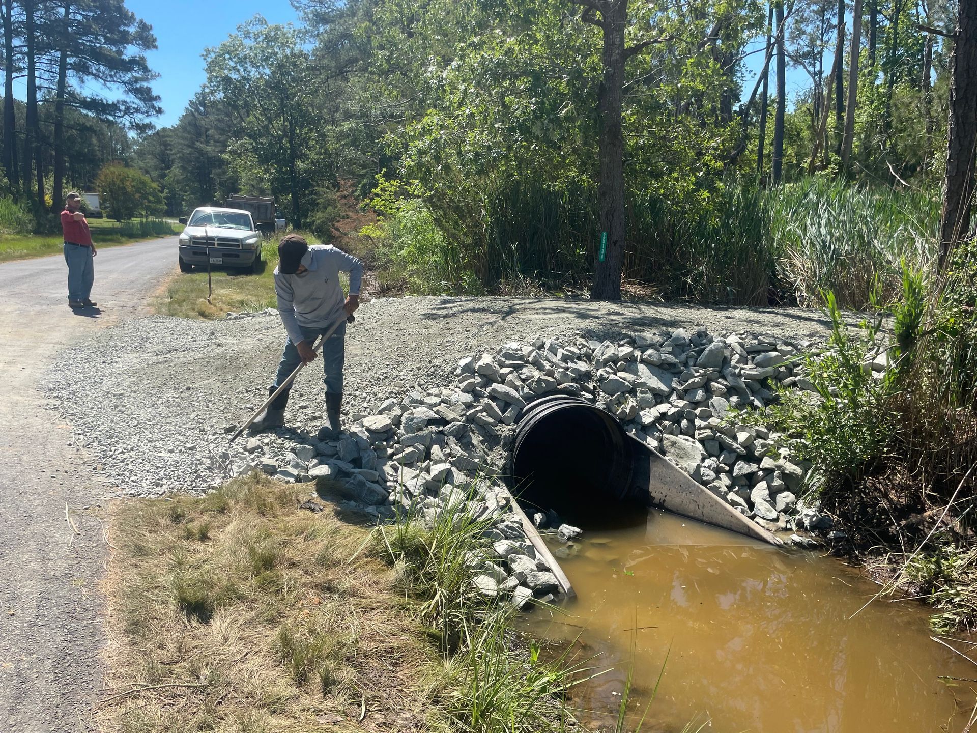 Person inspecting culvert on gravel road beside muddy creek, with a parked truck and trees nearby