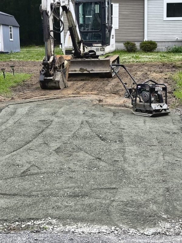 Excavator and plate compactor smoothing a gravel driveway beside a house.