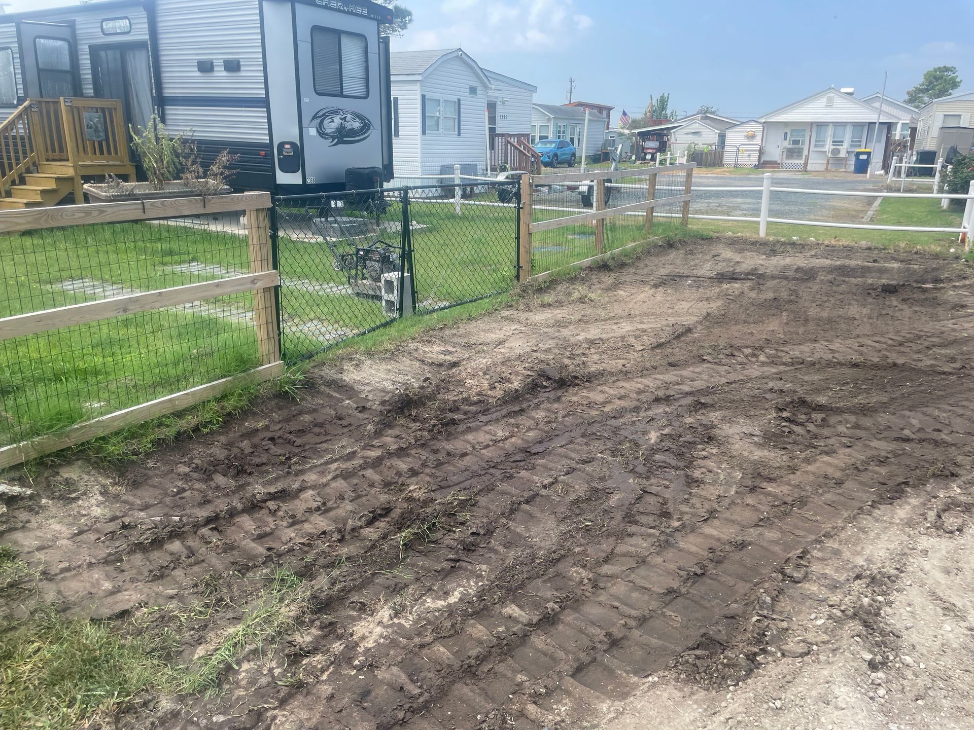 Dirt road beside a fenced grassy lot with parked RVs and houses in the background