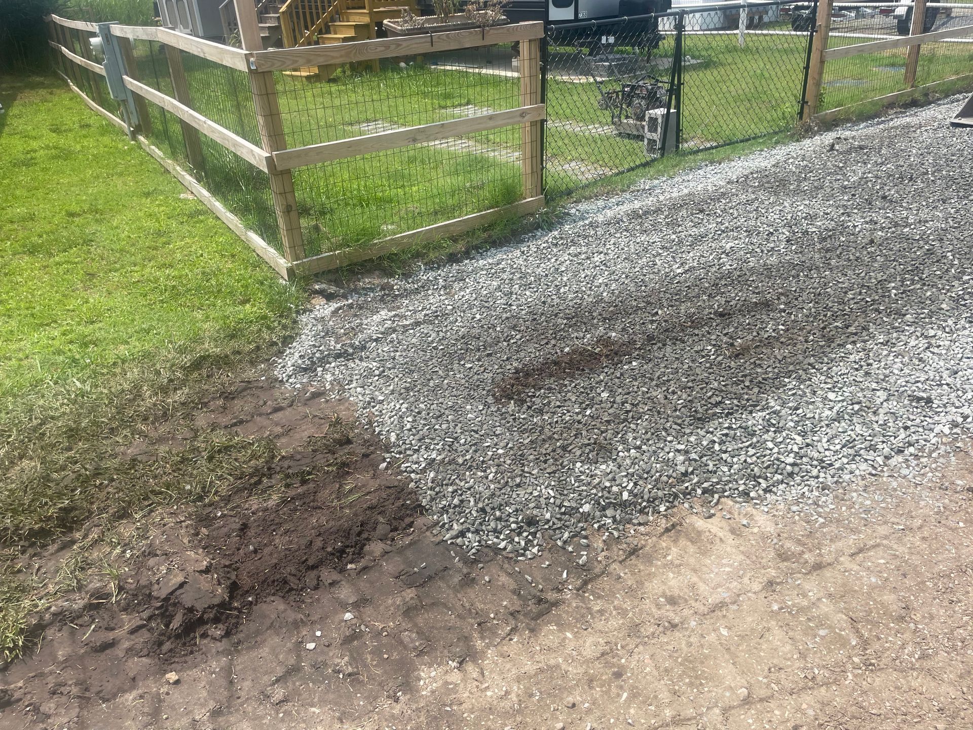 Gravel driveway beside muddy ground and a wooden fence along a grassy yard