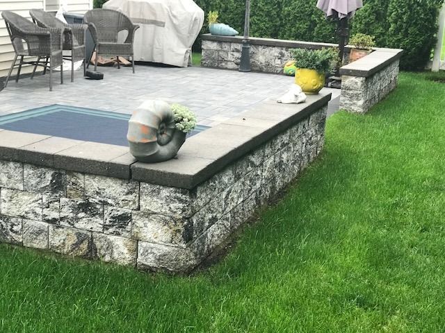 A stone wall surrounds a patio with a grill and chairs.