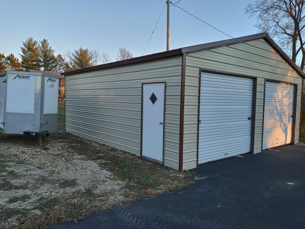 Tan metal two-car garage with a brown roof and trim, white doors, and a trailer parked nearby.