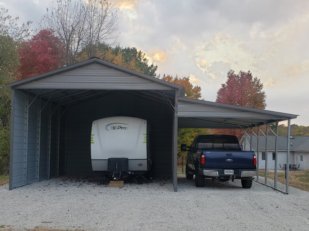 Gray metal carport sheltering a white camper and a blue pickup truck on a gravel surface. Trees in background.