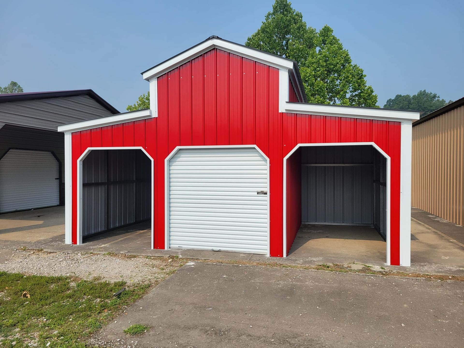 Red and white barn-style shed with a garage door and two open bays, on a gravel surface.