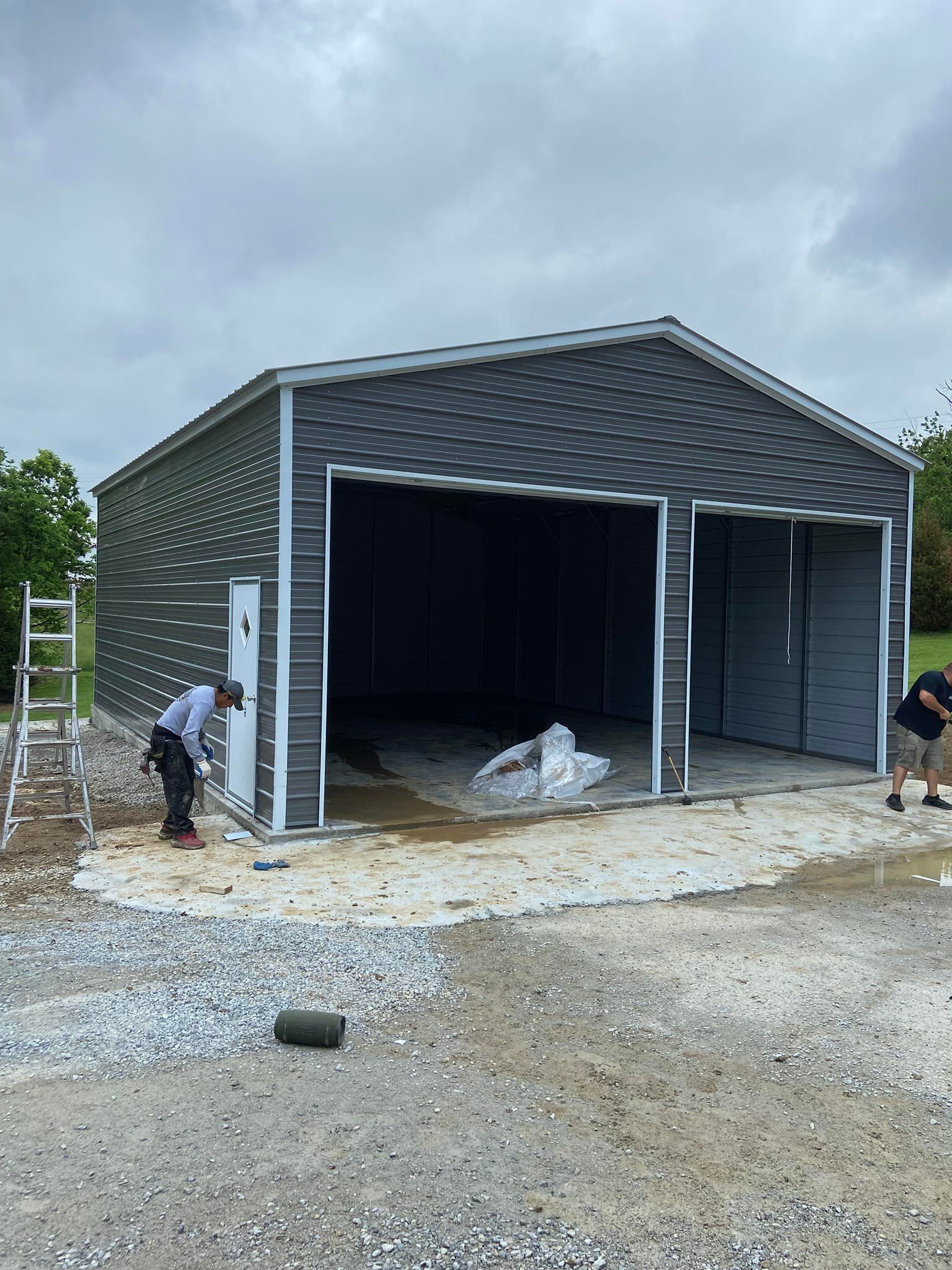Two-door metal shed under construction with two workers present on gravel. Gray walls and roof.