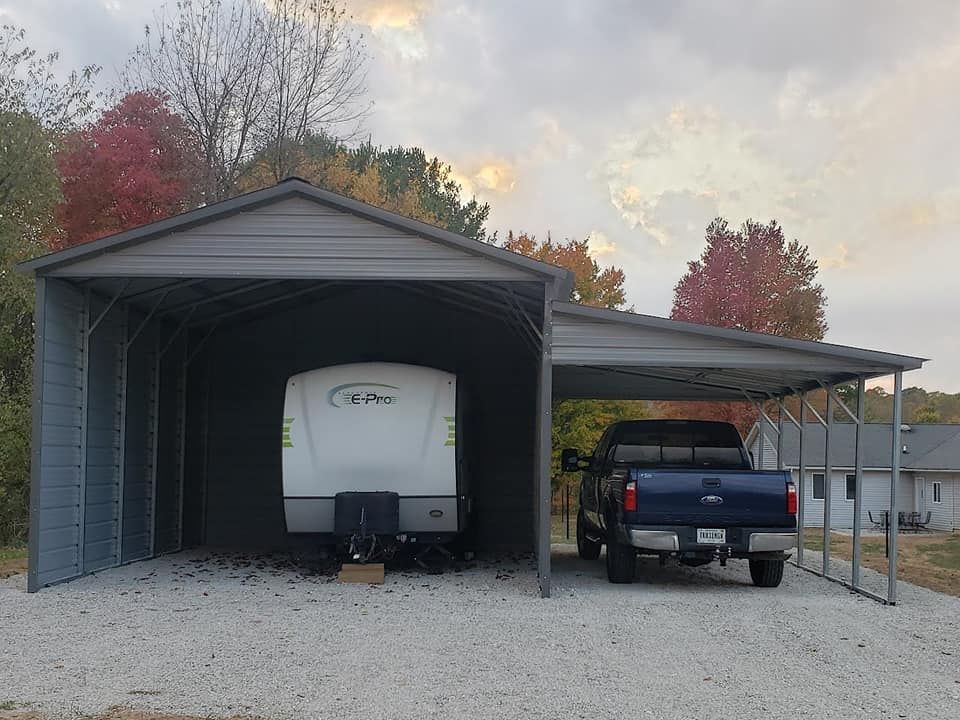 A metal carport shelters a camper and a blue pickup truck on a gravel driveway, with fall foliage in the background.