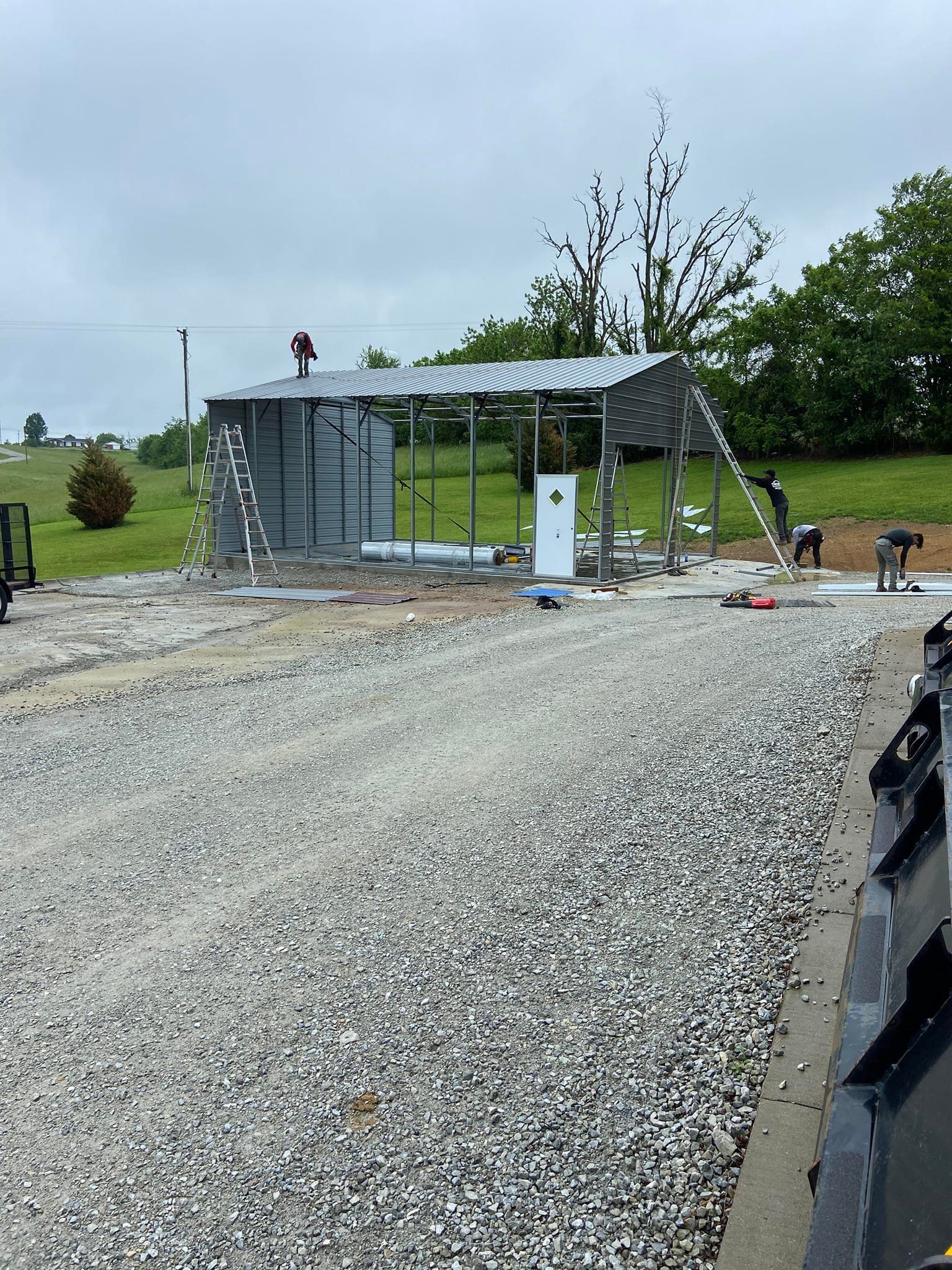 Construction of a metal shed with gravel driveway under overcast sky. Workers on roof.