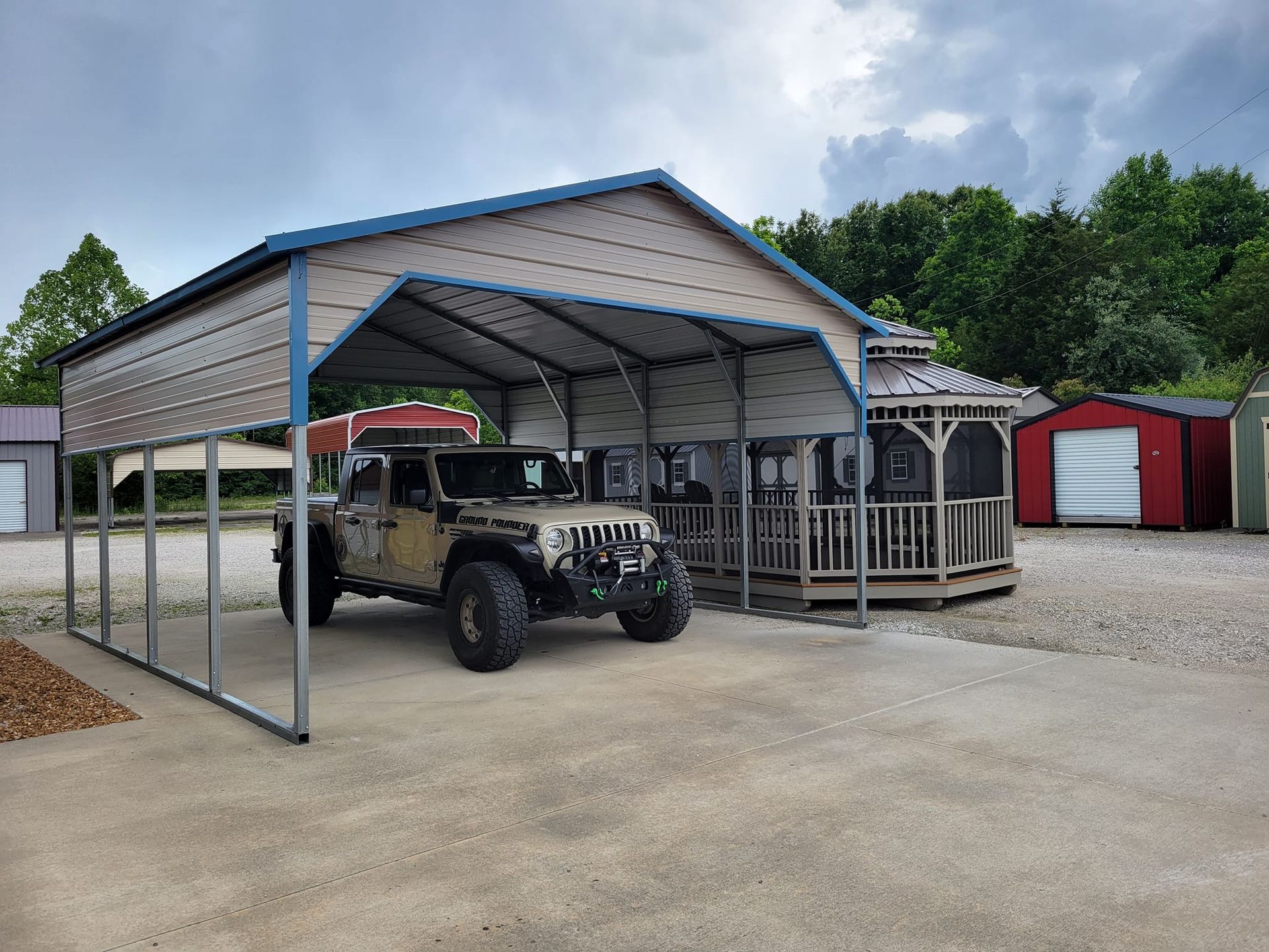 Tan Jeep parked under a metal carport next to a gazebo and storage sheds on a gravel lot.