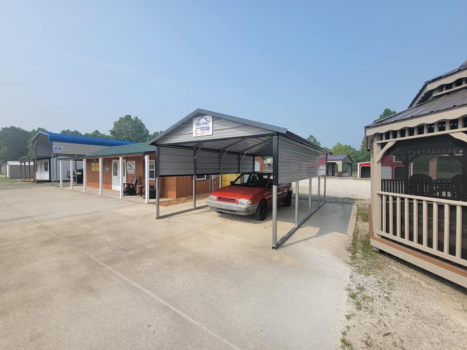 A red car parked under a metal carport. Several small buildings are visible in a lot under a clear sky.