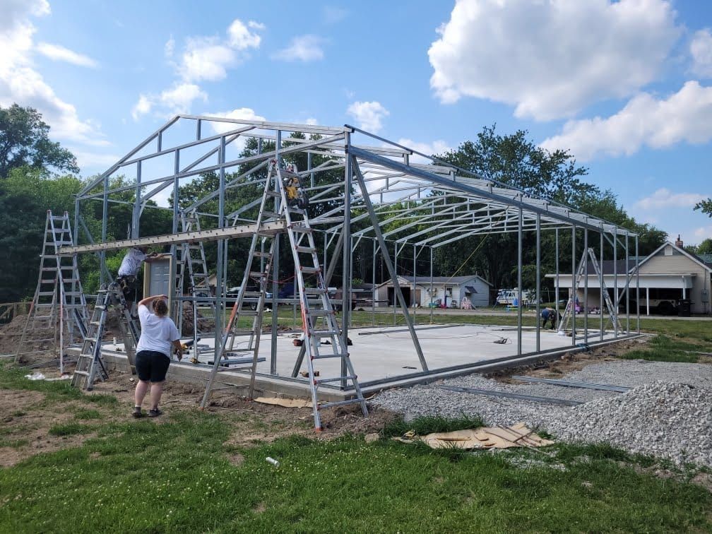 Metal frame building under construction, workers on ladders, concrete base, sunny day.