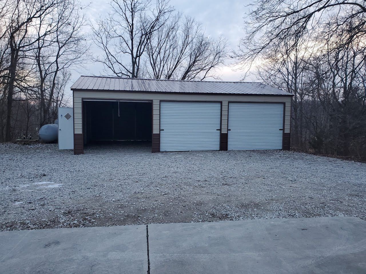 Three-bay garage with metal roof, gravel driveway. Two closed garage doors, one open bay. Gray and tan colors.