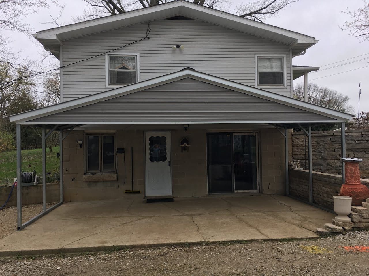 Two-story house with a covered patio. Gray siding, front door, windows. Concrete patio, metal supports.