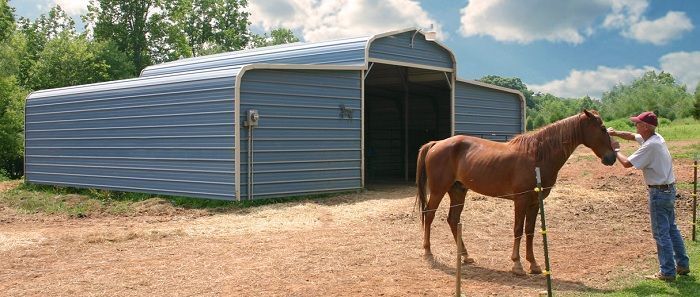 A person pets a brown horse near a blue metal barn on a sunny day.