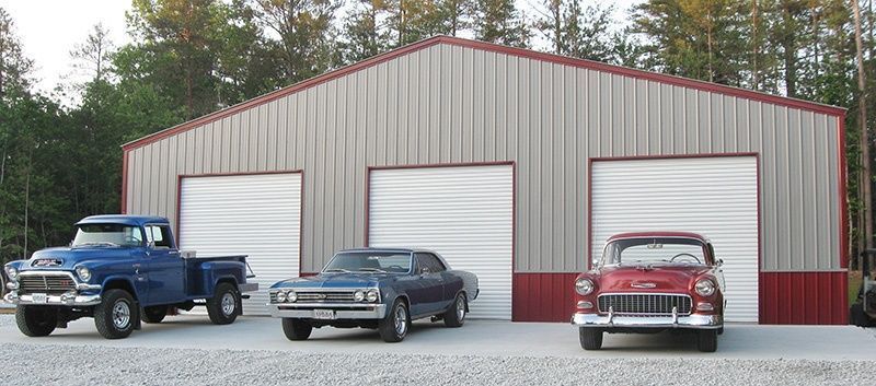 Three classic cars parked in front of a metal garage with three bays.
