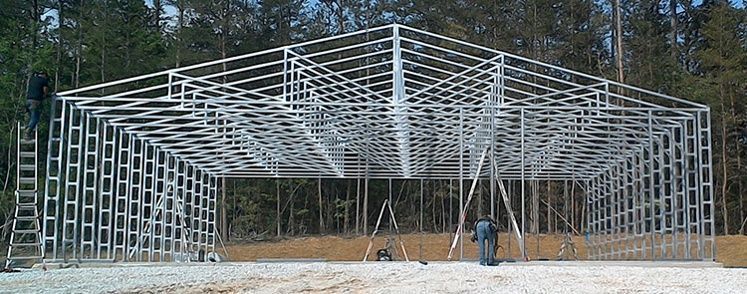 Metal frame of a building under construction, set in a wooded area. A worker stands below.