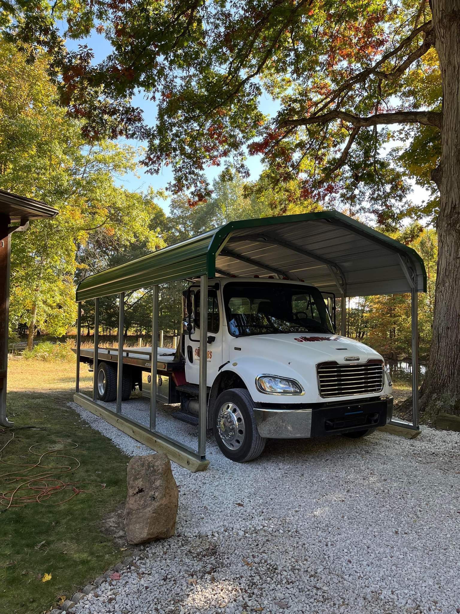White truck under a metal carport in a gravel driveway, surrounded by trees.