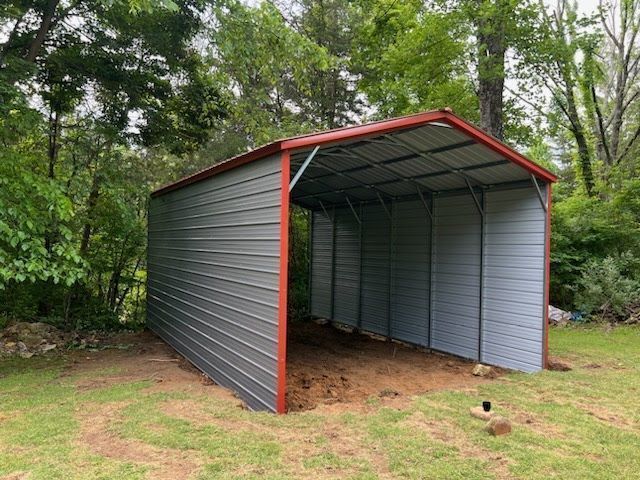 Gray metal carport with red trim, open-sided, sits on a grassy area, trees in background.