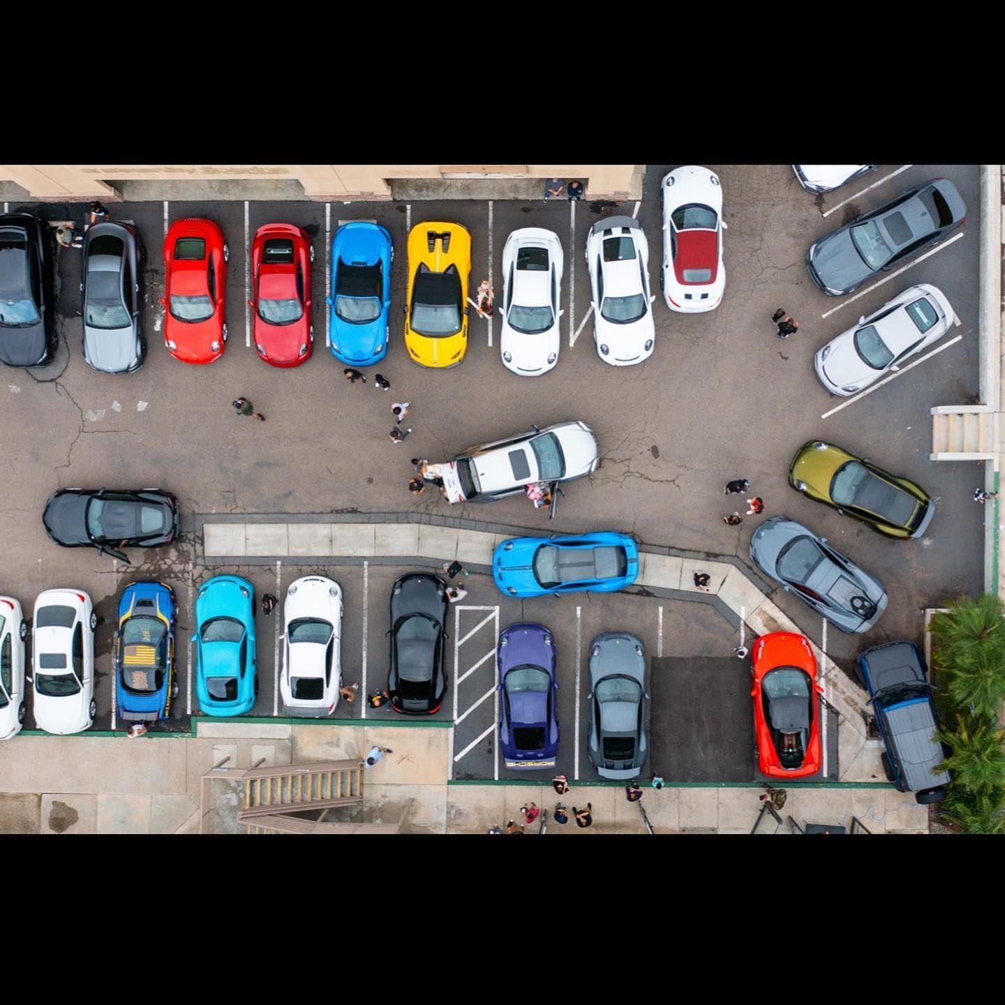 Overhead view of a parking lot filled with colorful sports cars. People mill about the vehicles.