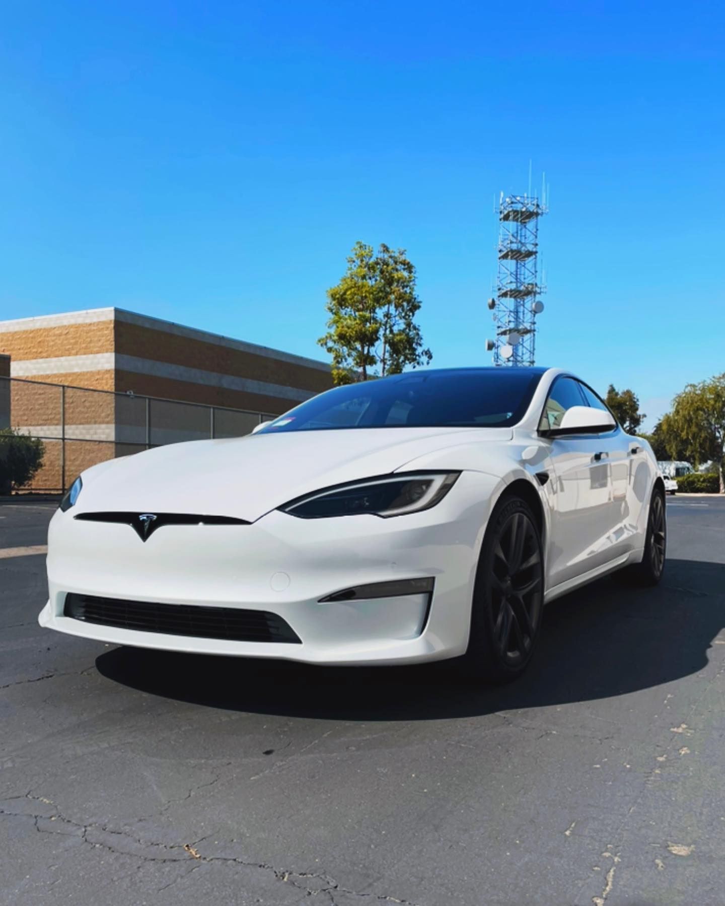 White Tesla car parked in a paved lot, with a blue sky and industrial buildings in the background.