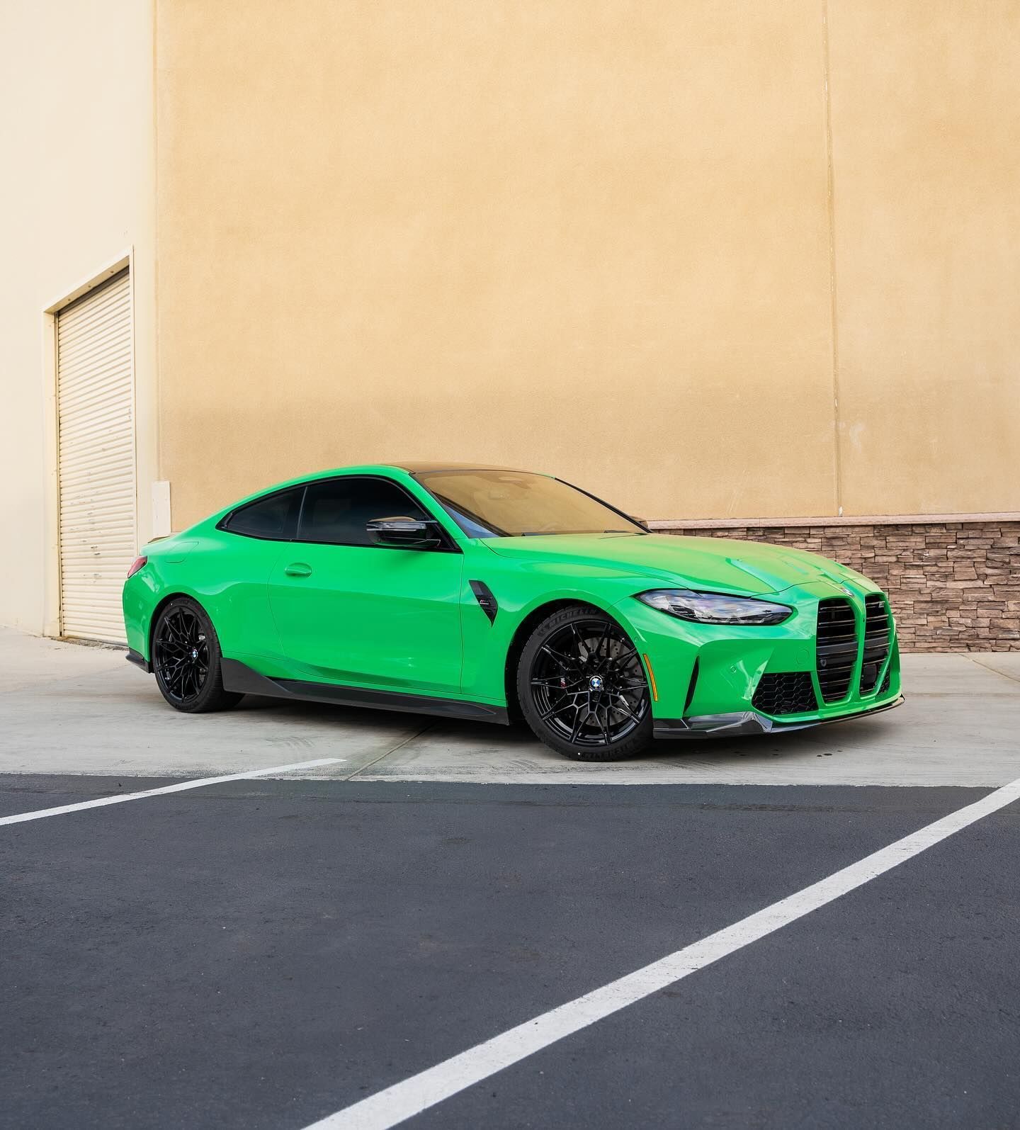 Green BMW M4 coupe parked on asphalt, near a light yellow building. Black wheels.