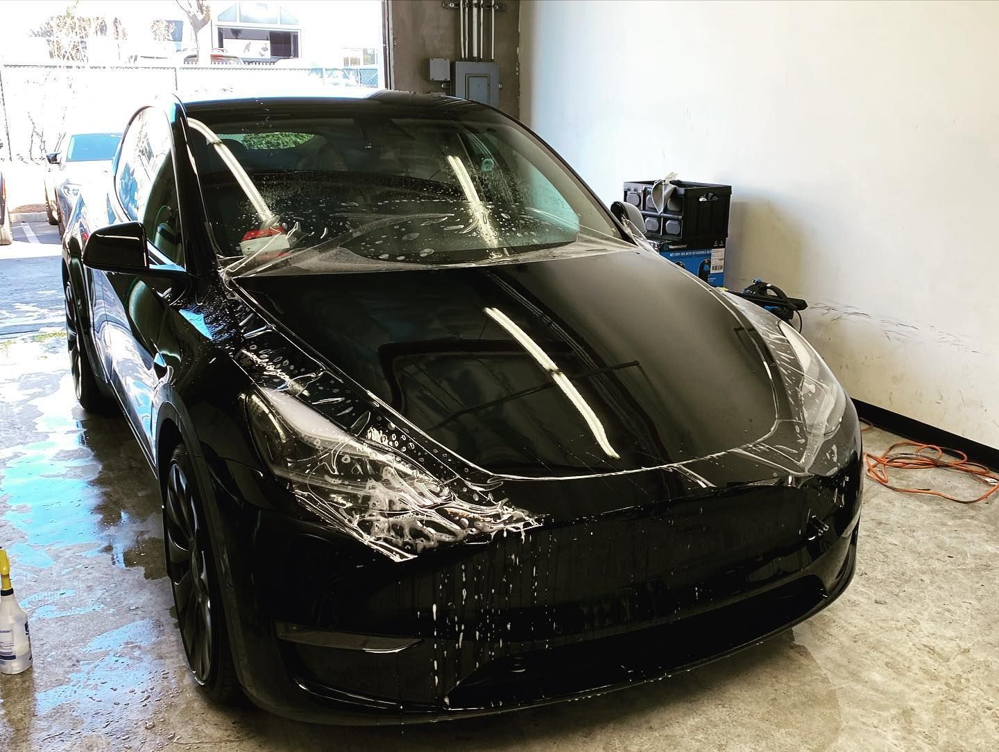 Black Tesla Model Y being washed, covered in soap suds, in a garage.