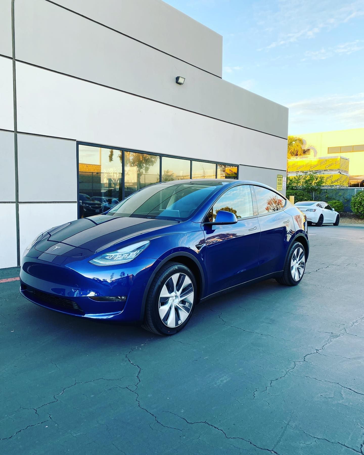 Blue Tesla Model Y parked in front of a modern building with blue sky.