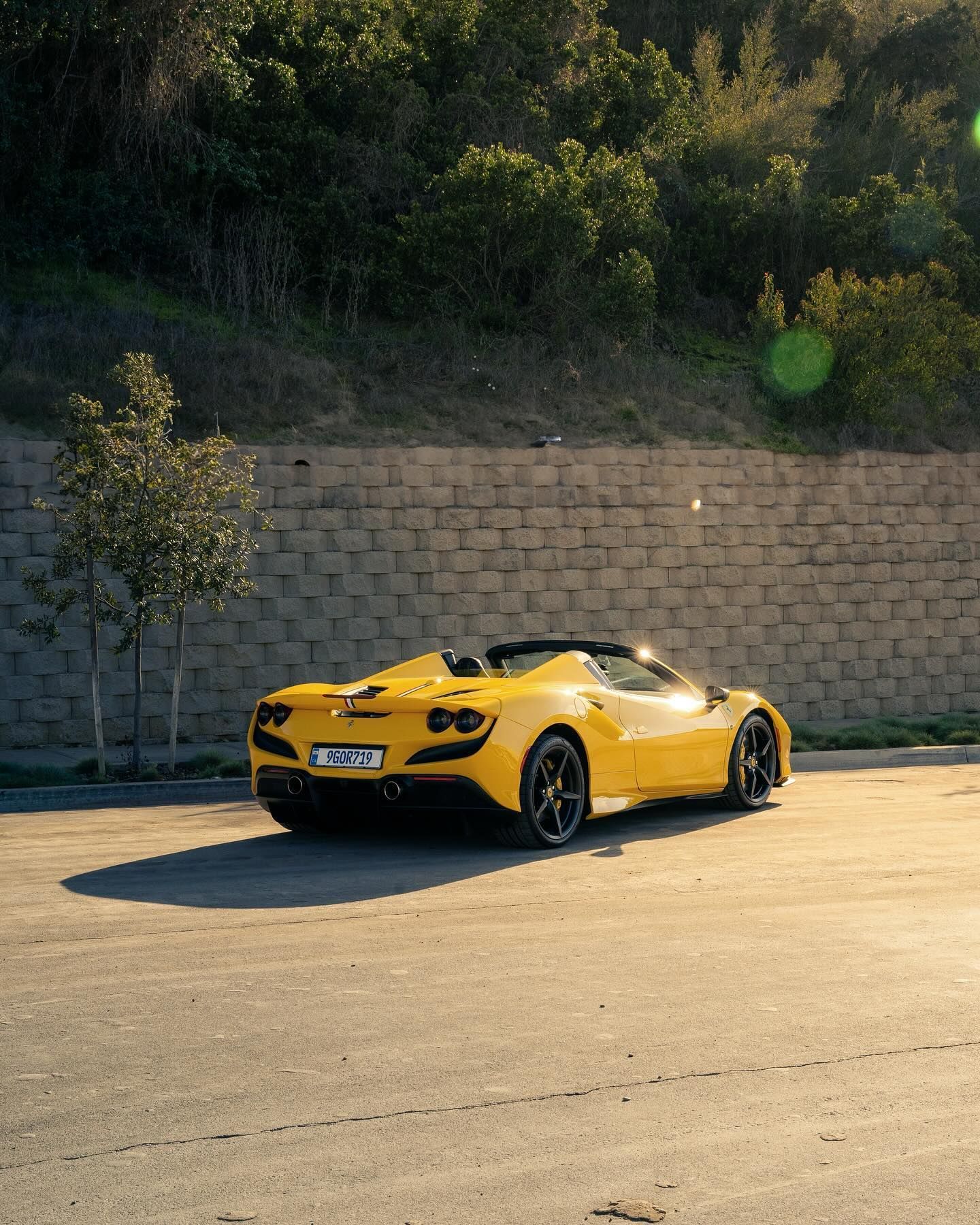 Yellow Ferrari convertible parked on asphalt in front of a stone wall and trees.