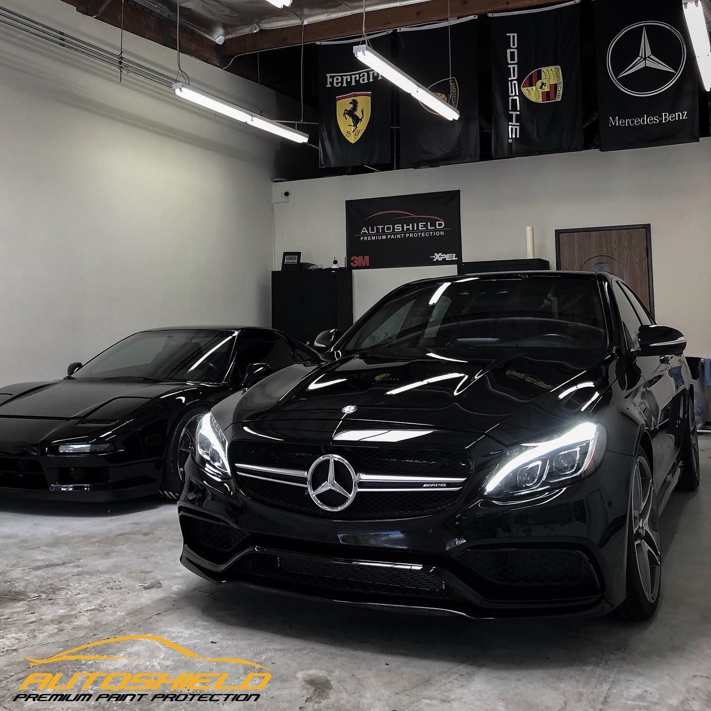 Two black cars, a Mercedes and an older sports car, in a garage with car brand flags.