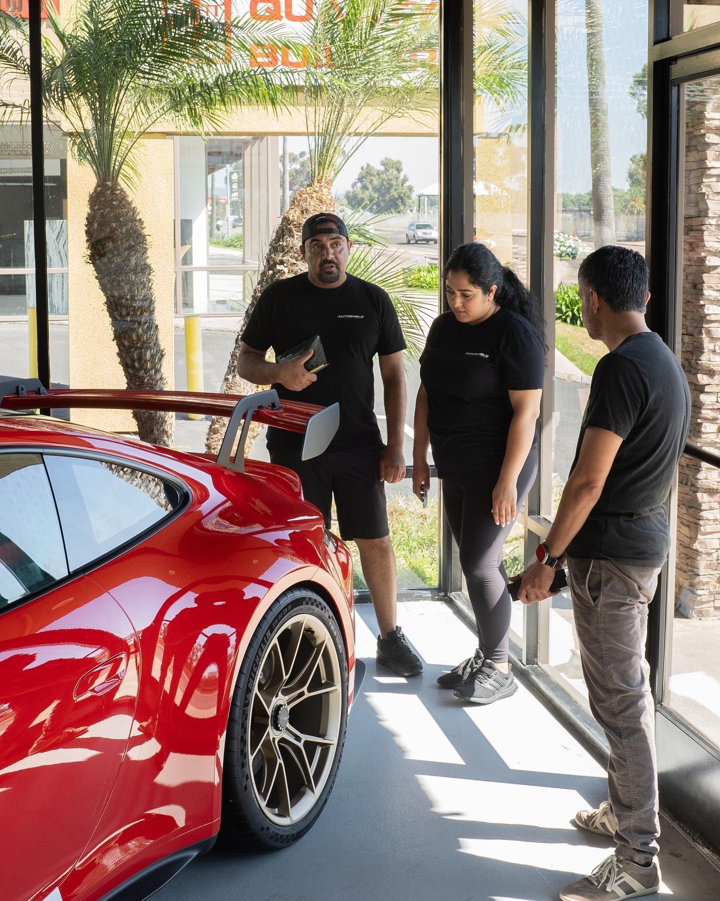 Three people examining a red sports car in a showroom. Bright lighting, full car in frame.