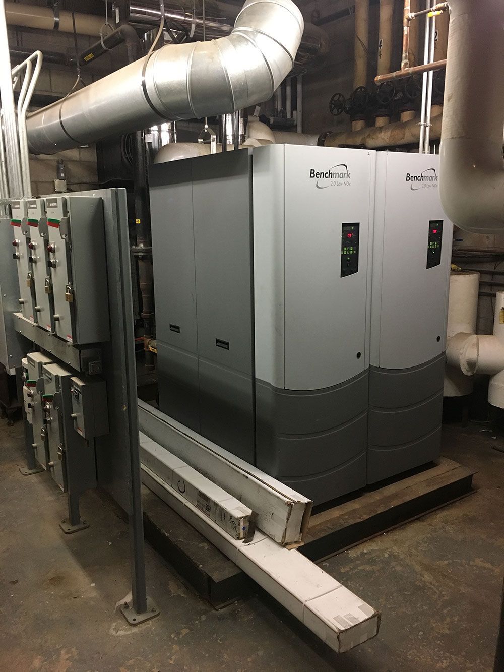 Gray industrial chiller unit in a mechanical room with pipes and electrical boxes.