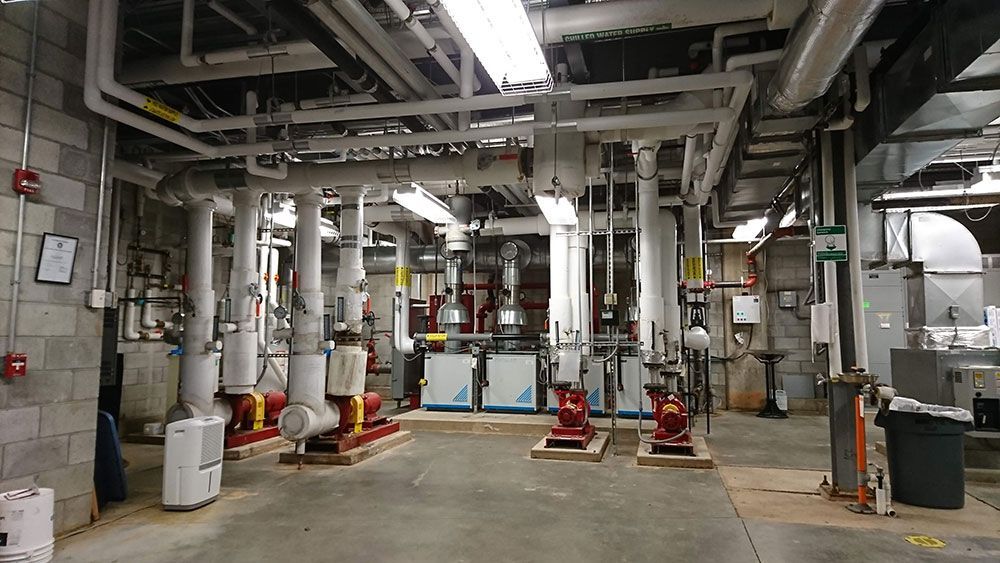 An industrial mechanical room with pipes, pumps, and other equipment against concrete walls and floor.