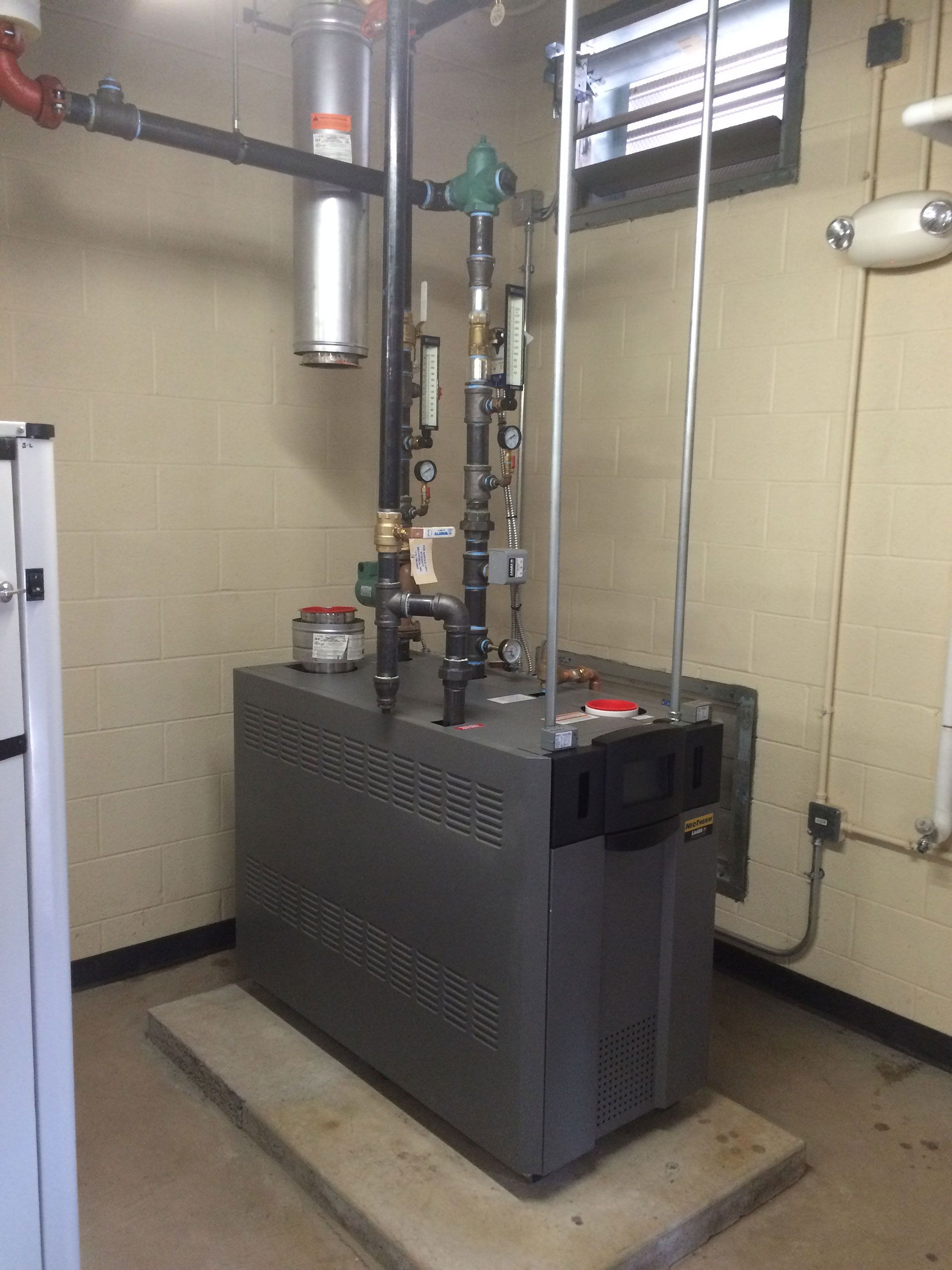 Boiler with pipes and gauges in a utility room. Gray metal unit with black pipes against beige walls.