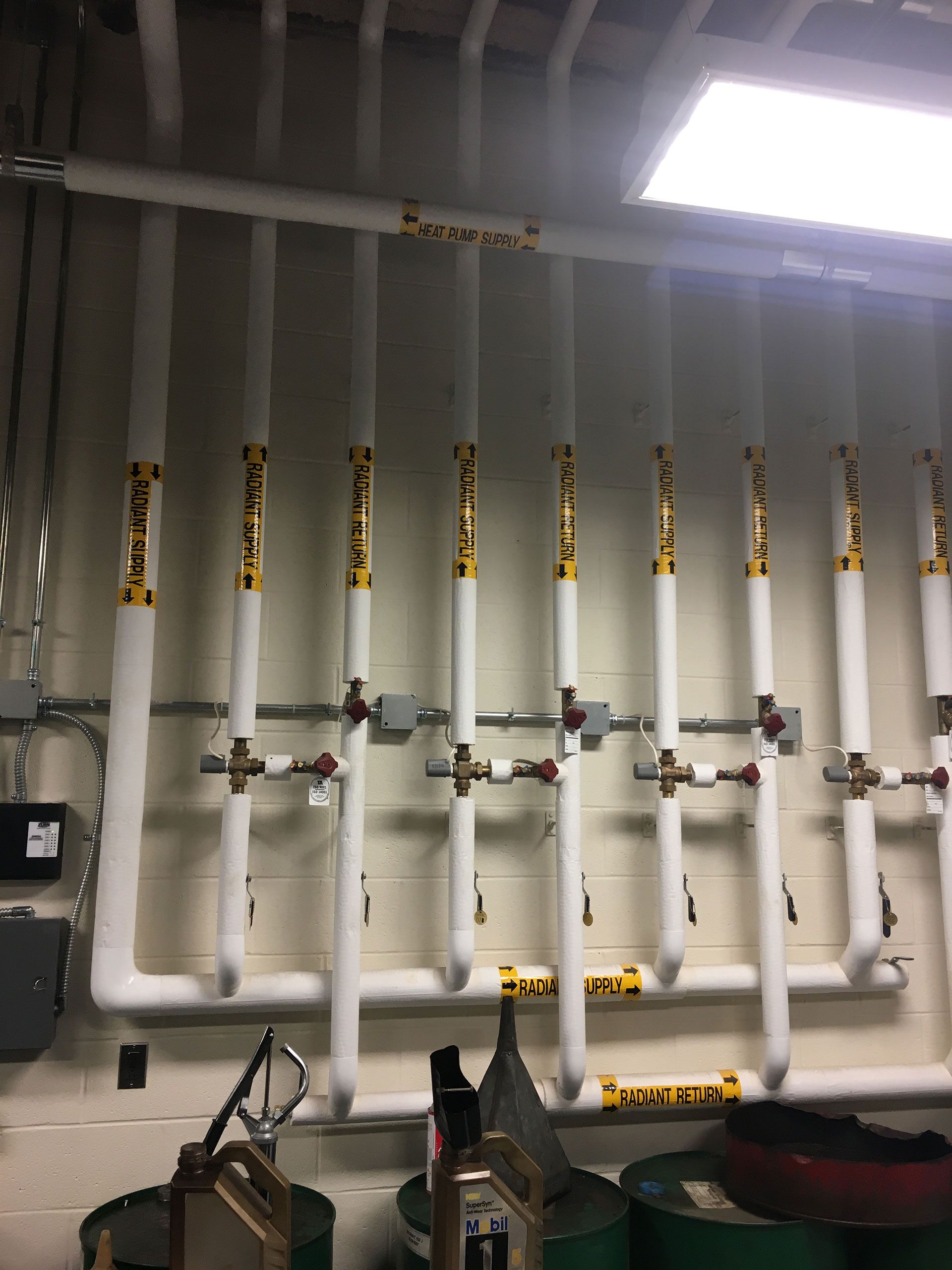 White insulated pipes run vertically along a wall with valves and yellow labels.