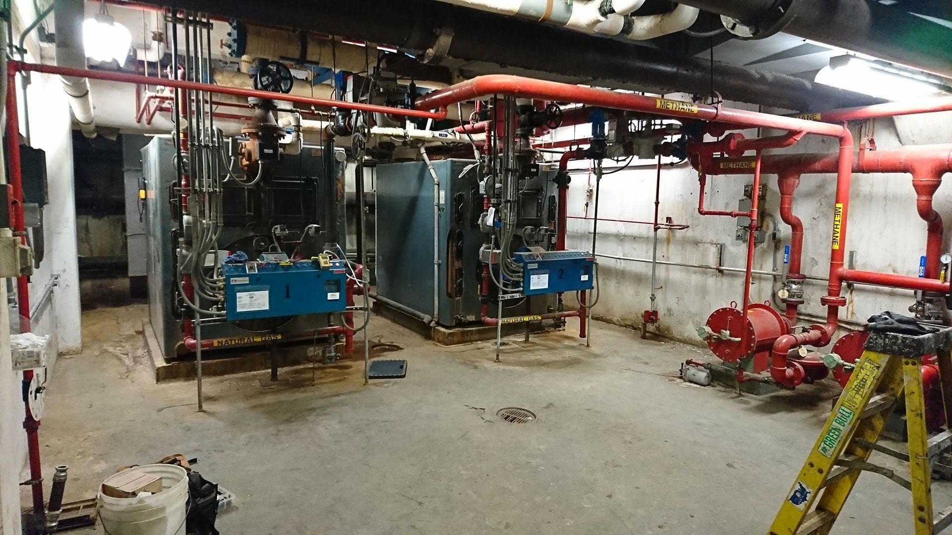 Large industrial equipment room with red pipes, two large metal boxes, and a yellow ladder.