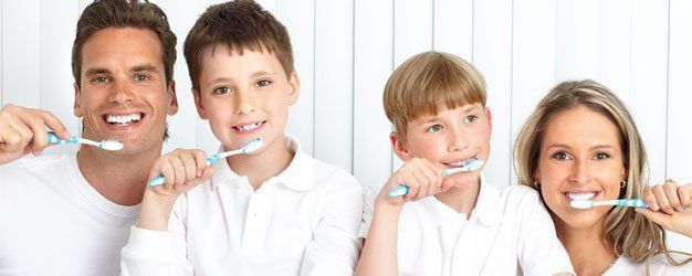 Family brushing teeth together in a bright bathroom, smiling at the camera.