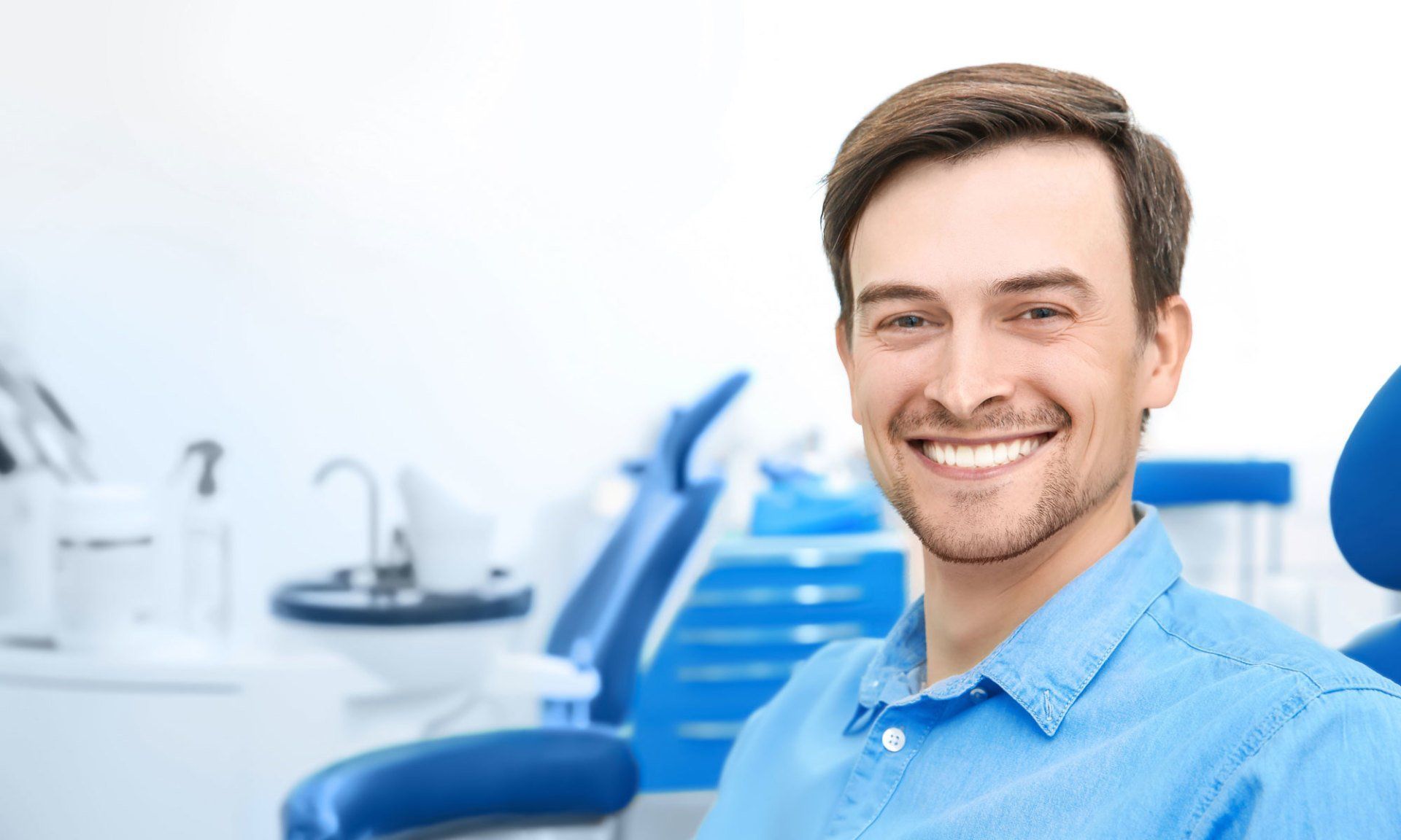 Smiling man in blue shirt at a bright dental clinic with a chair and dental tools behind him