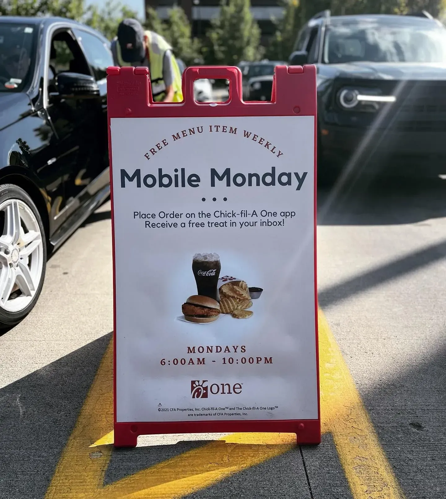 Red sidewalk sign promoting Mobile Monday at a café, with a black car parked behind it.