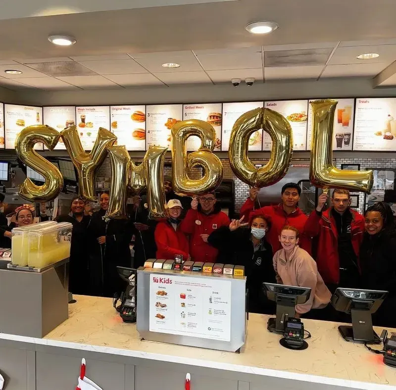 Restaurant staff posing behind a counter with gold balloon letters spelling “SYMBOL.”