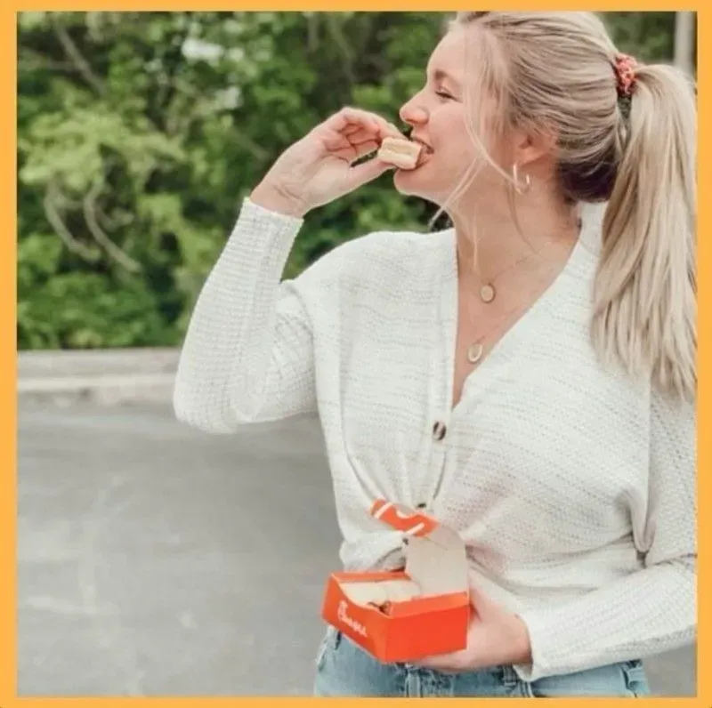 Woman outdoors eating a snack, holding an orange box, wearing a white cardigan and jeans.