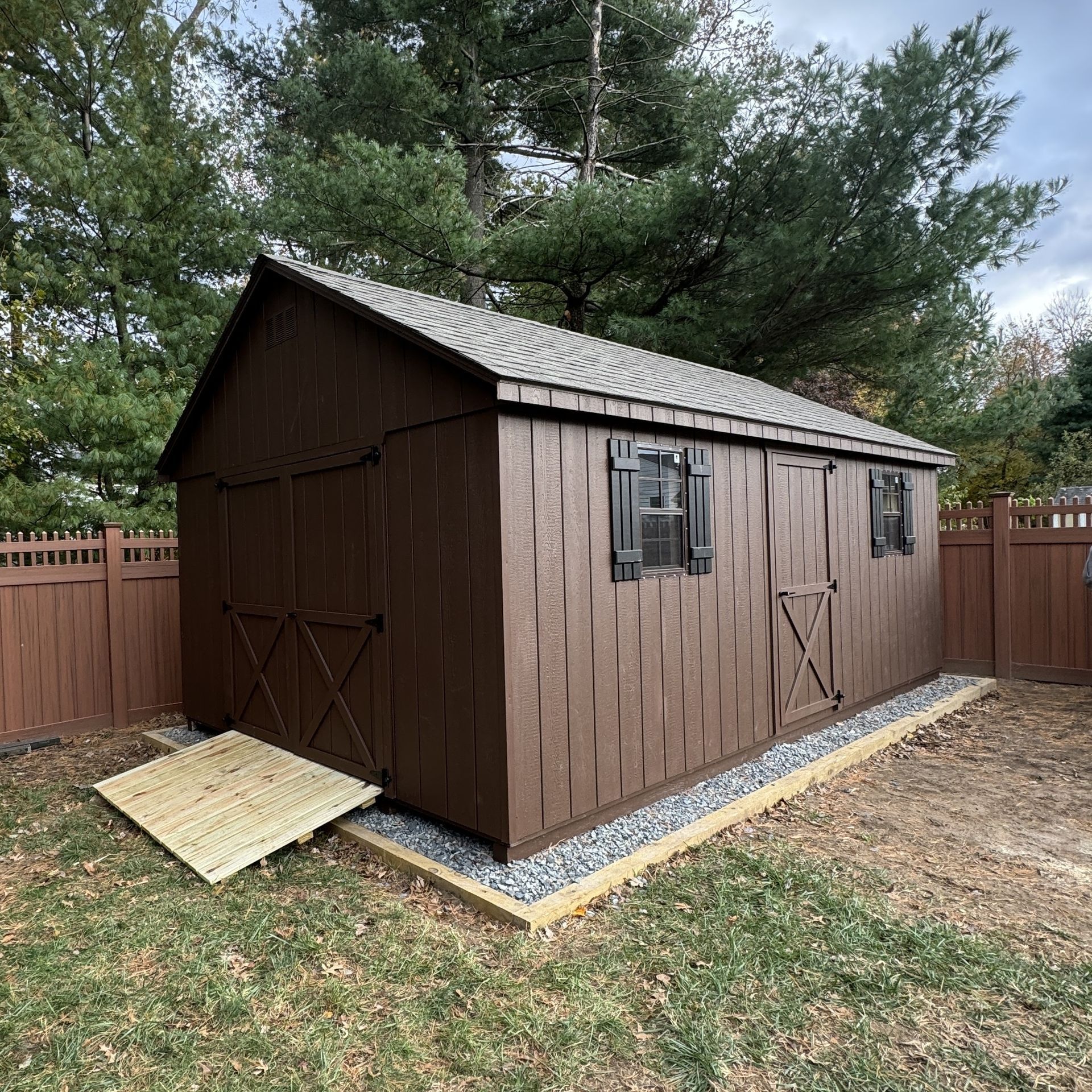 Brown backyard shed with double doors, side window, and ramp, surrounded by a wooden fence and trees.