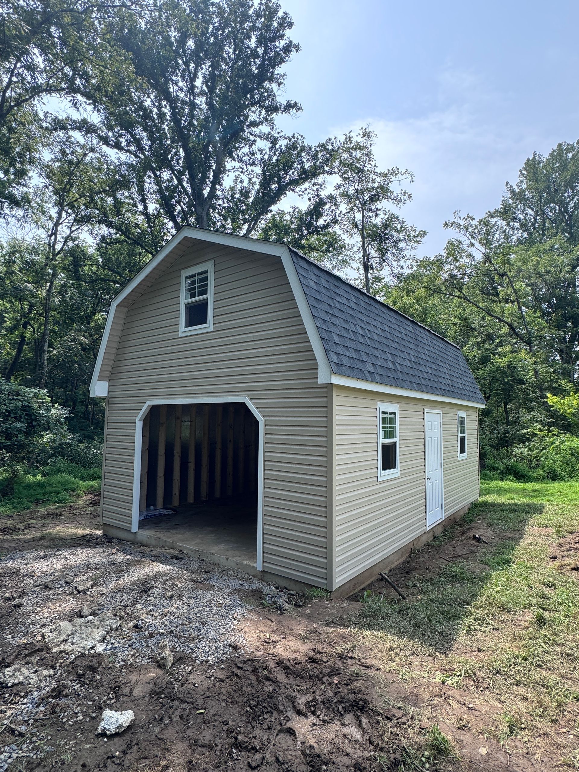 Small beige shed with open garage-style door, gravel ramp, and white side door in a wooded yard.