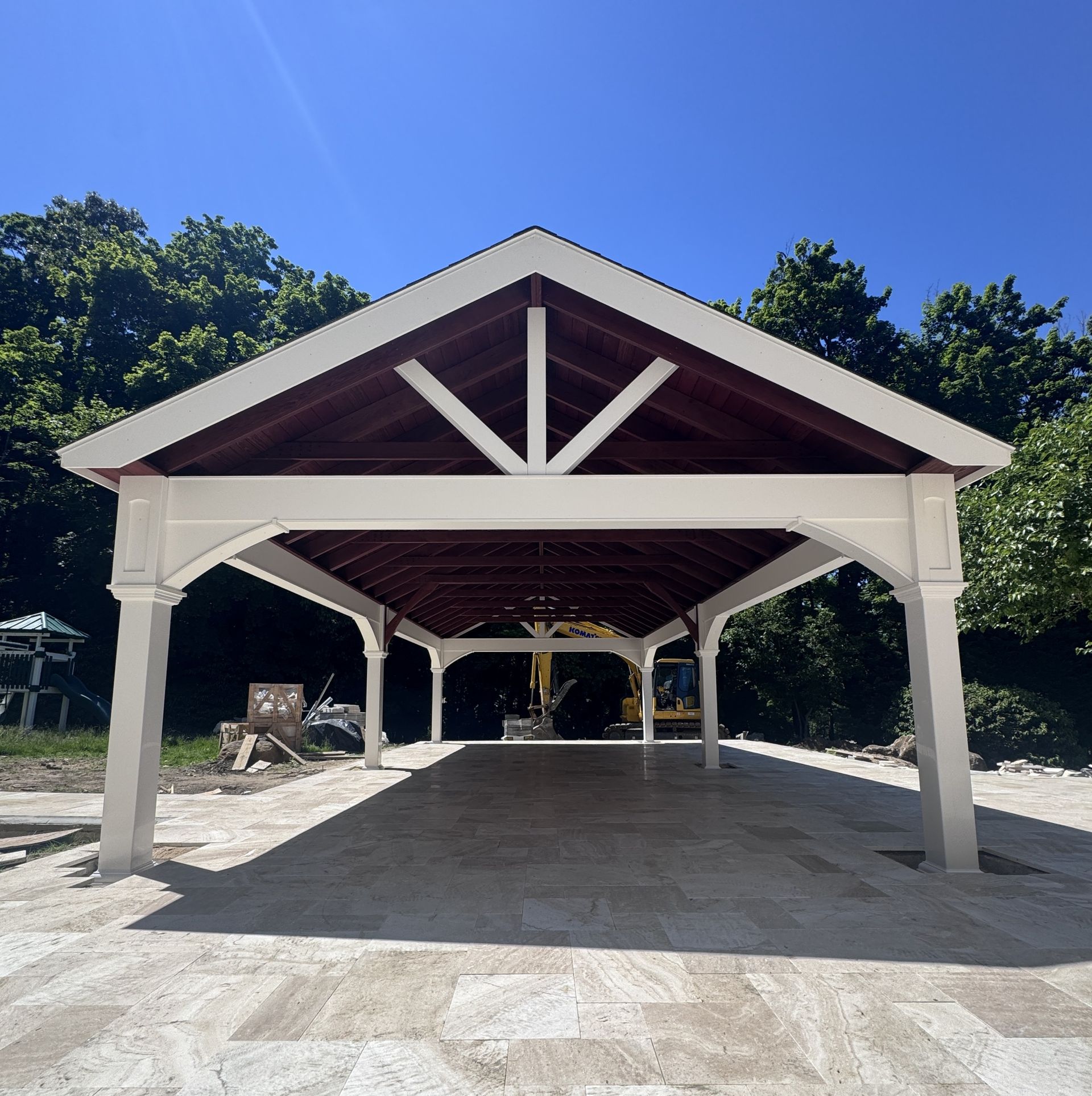 Open-air pavilion with white posts and dark red roof under a blue sky, surrounded by trees